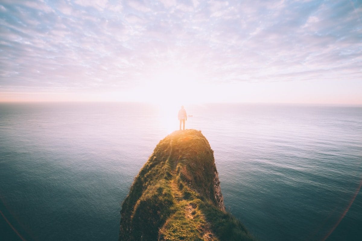 Person standing at the edge of a cliff overlooking the ocean at sunrise — representing purpose and direction